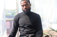 Man wearing a black turtleneck sweater sitting in a room with large windows.