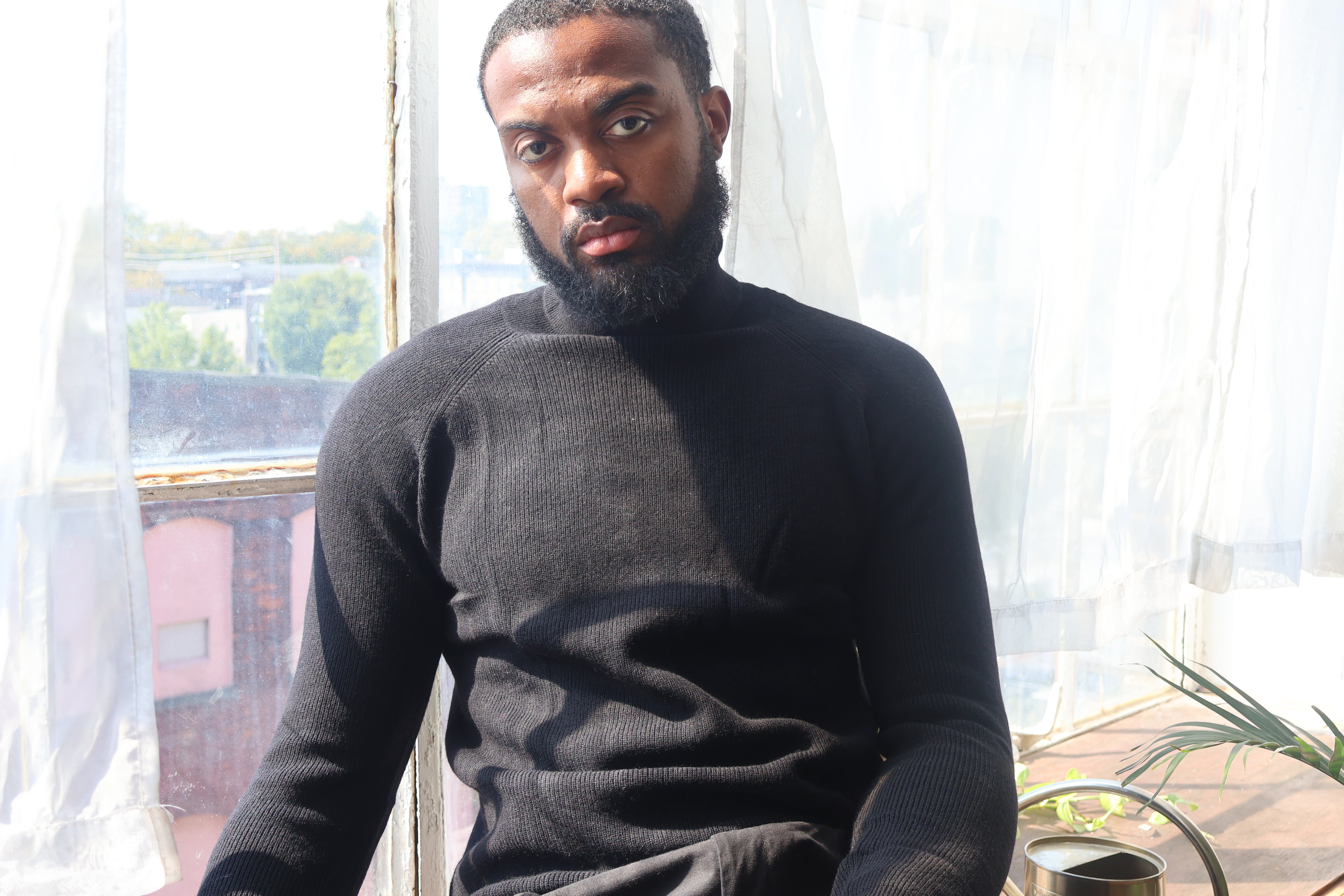 Man wearing a black turtleneck sweater sitting in a room with large windows.